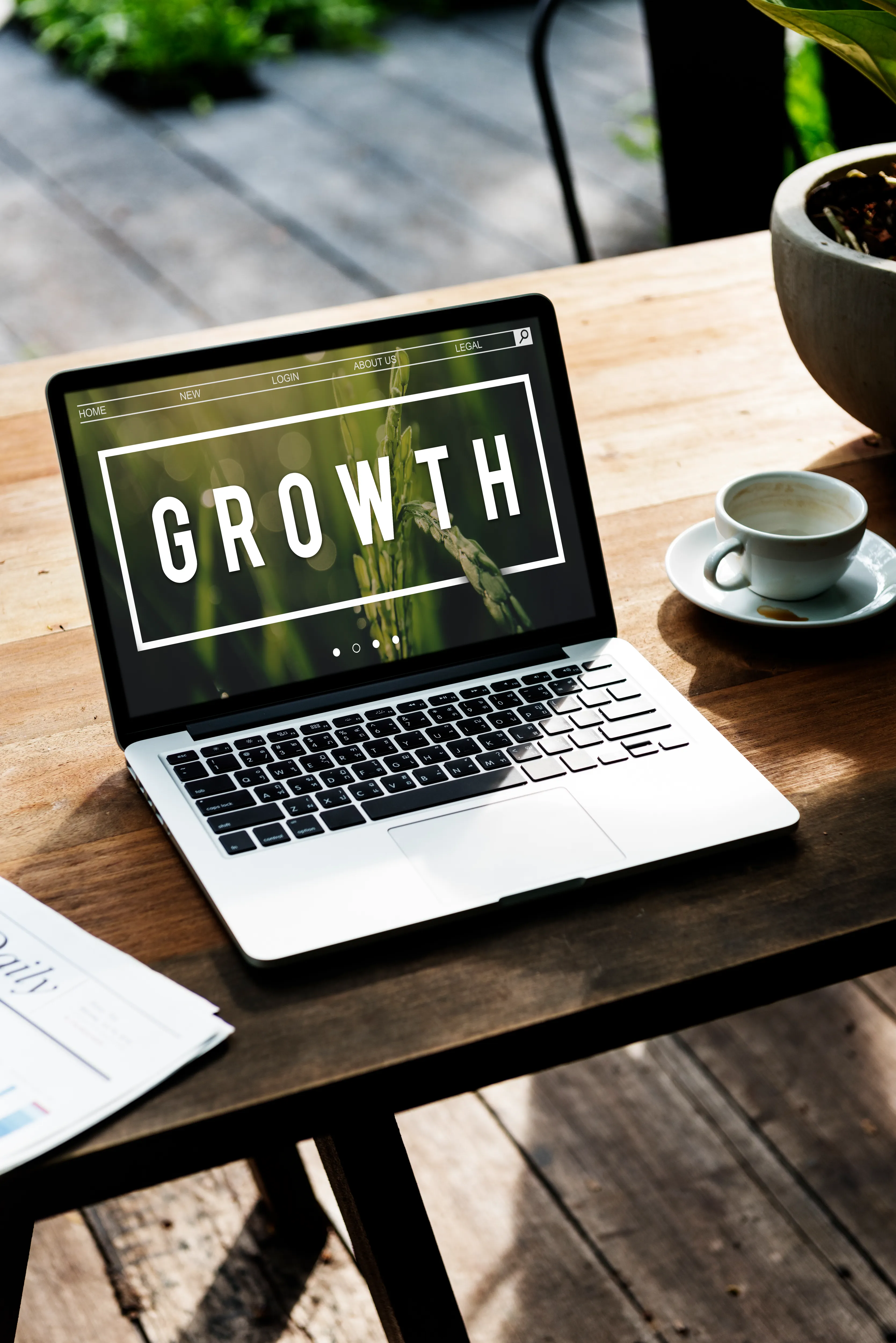 Laptop displaying GROWTH on a wooden desk beside a teacup and newspaper
